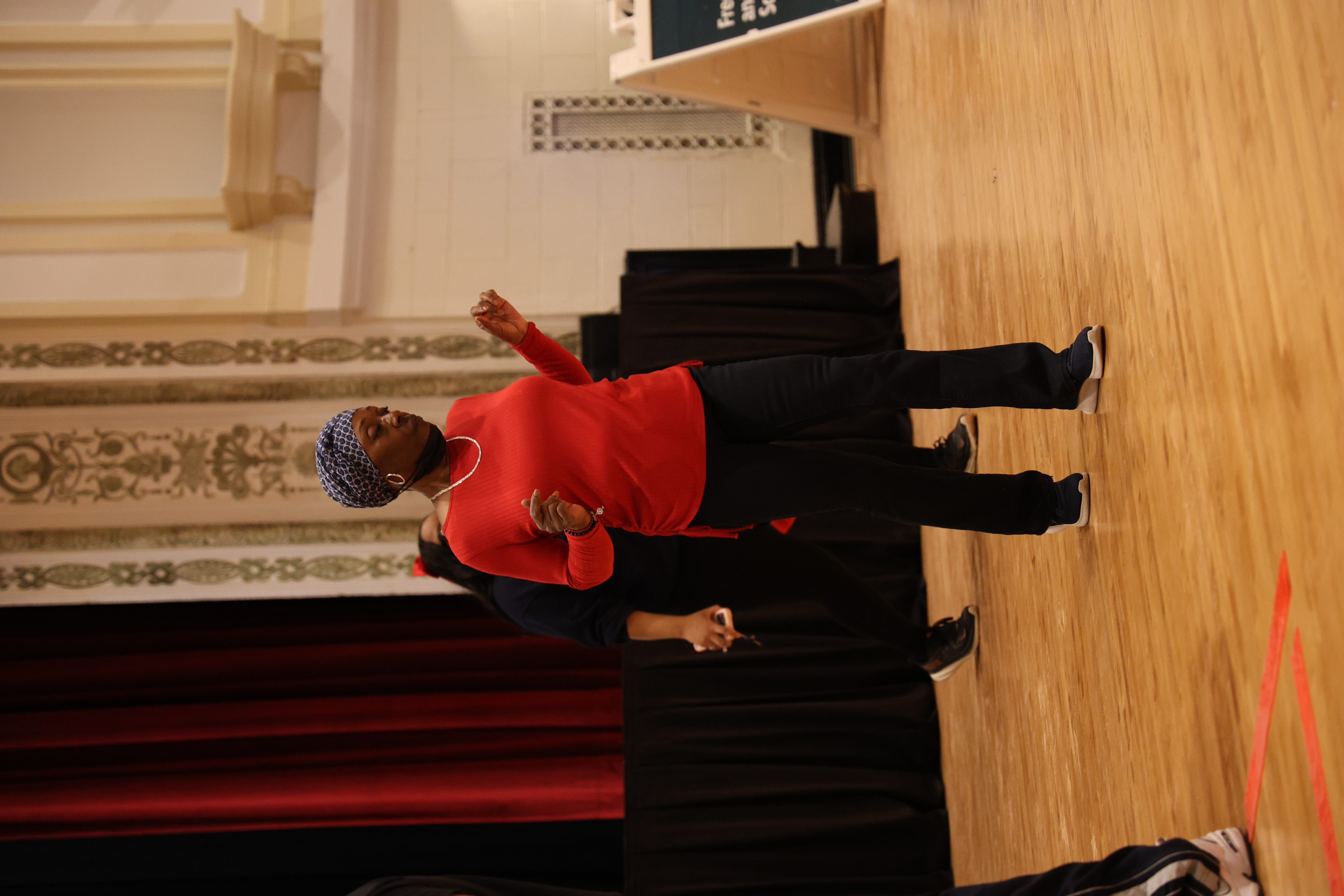 African American woman in red shirt and patterned headwrap dances on a wooden floor.